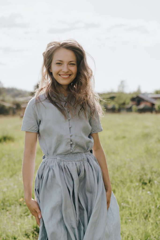 A young woman in a blue dress smiles brightly in a lush green field, epitomizing carefree summer vibes.