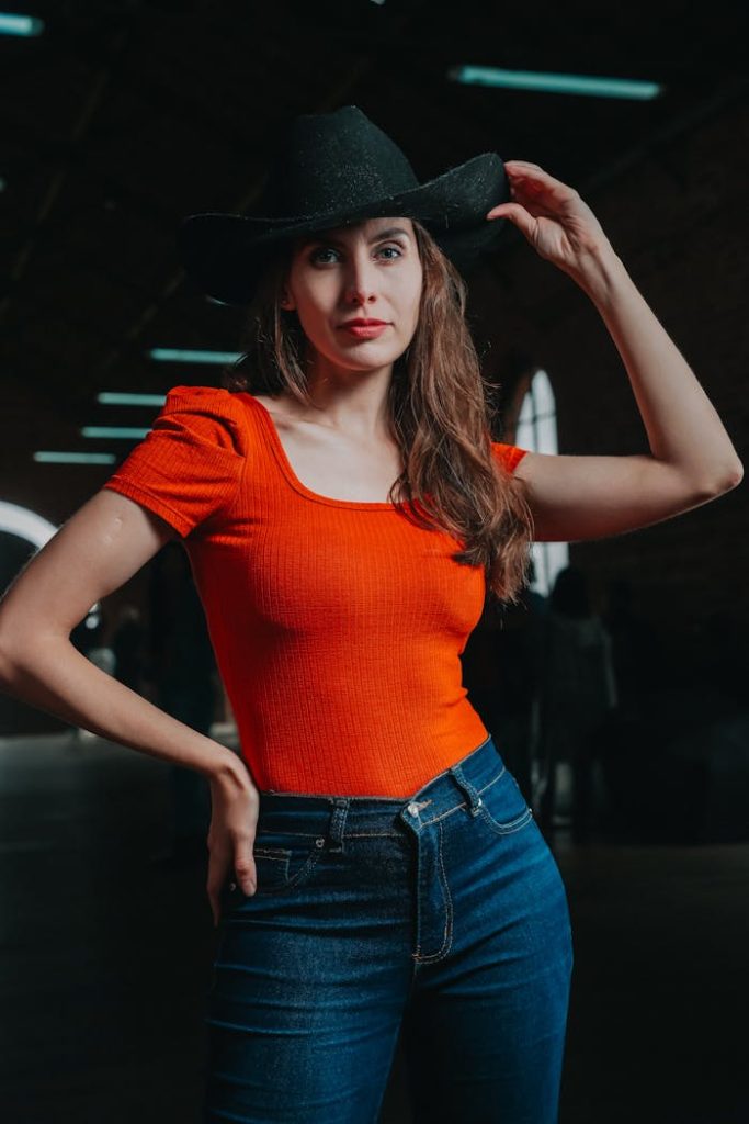 Fashionable woman wearing a cowboy hat and red top in a dimly lit indoor setting, posing confidently.